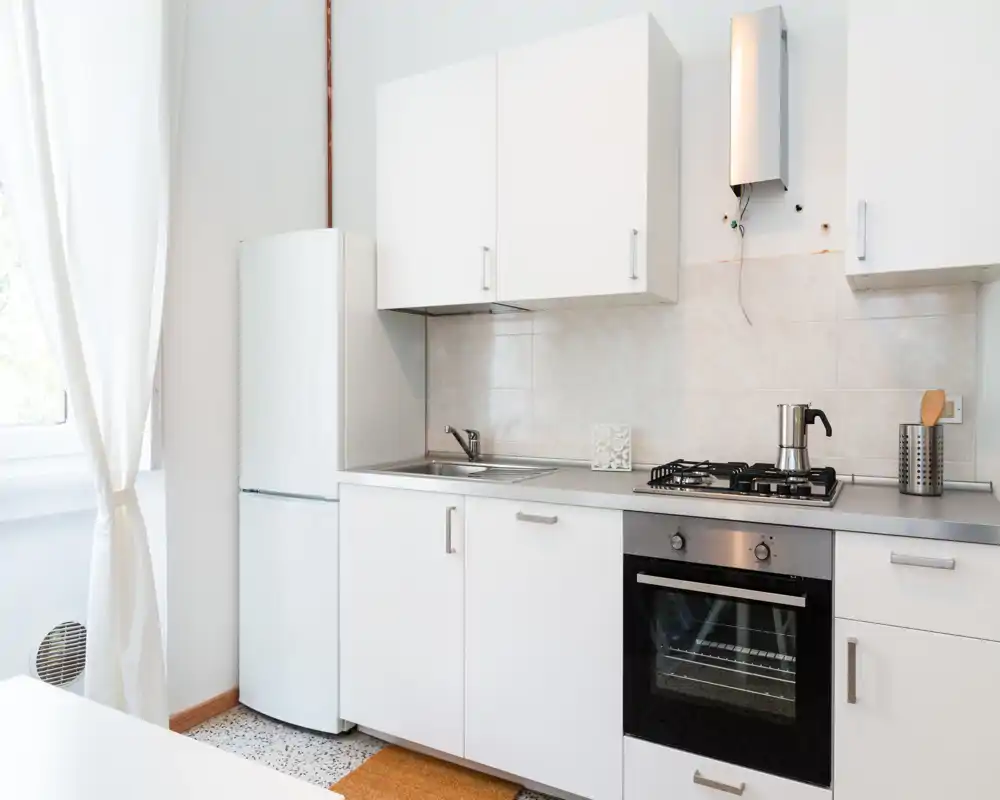 Cucina luminosa e minimal con pensili bianchi, forno integrato e piano a gas, frigorifero e lavello in acciaio. La luce naturale dalla finestra e la decorazione essenziale creano un ambiente pulito e arioso.