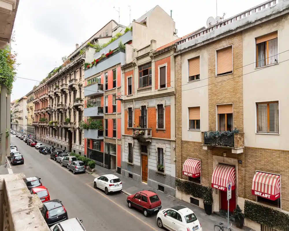 Vista di una tranquilla strada cittadina e delle facciate dei palazzi vista dal balcone; auto parcheggiate lungo la carreggiata e tende di piccoli negozi visibili.