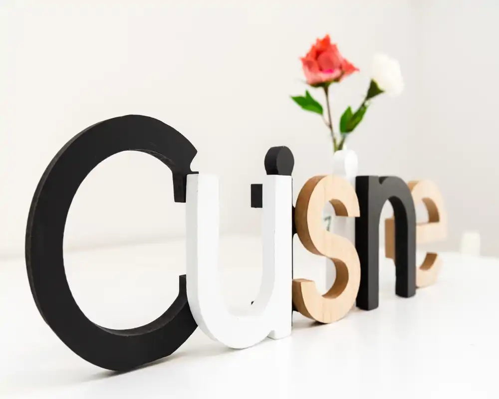 Primo piano decorativo di lettere in legno che formano 'Cusine' (probabilmente inteso come 'Cuisine' o 'Cucina') su una superficie bianca con un piccolo vaso e fiori sfocati sullo sfondo. L'immagine mostra tipografia e oggetti di arredo, non l'ambiente della stanza.