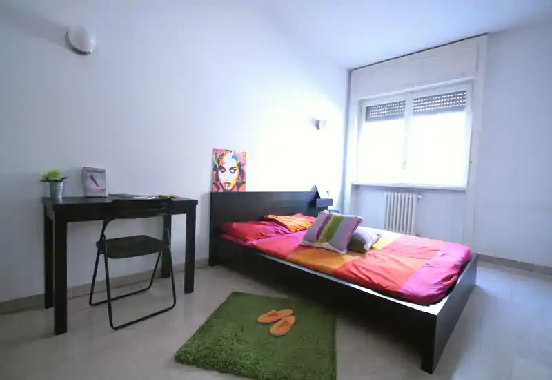 Dormitorio moderno y luminoso con cama baja de marco negro, ropa de cama y cojines coloridos, un pequeño escritorio y una ventana que aporta luz natural — ideal para una persona o estancias cortas.