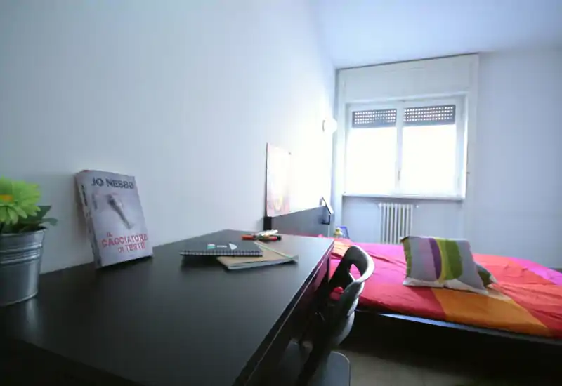 Dormitorio luminoso y acogedor con un escritorio sencillo en primer plano y una cama colorida junto a la ventana. La luz natural ilumina la habitación y el área de trabajo ordenada es ideal para alquileres cortos o estudiantes.