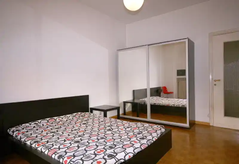 Dormitorio moderno y bien iluminado con cama doble, armario con puertas espejadas y mesita sencilla. Paredes neutras y ropa de cama estampada que crean un espacio limpio y acogedor.