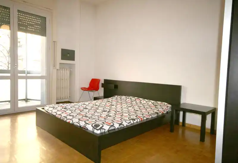 Dormitorio luminoso y minimalista con cama doble en estructura baja de madera oscura, suelo de parquet y puerta de balcón que aporta luz natural. Mesita sencilla y una silla roja como detalles funcionales.