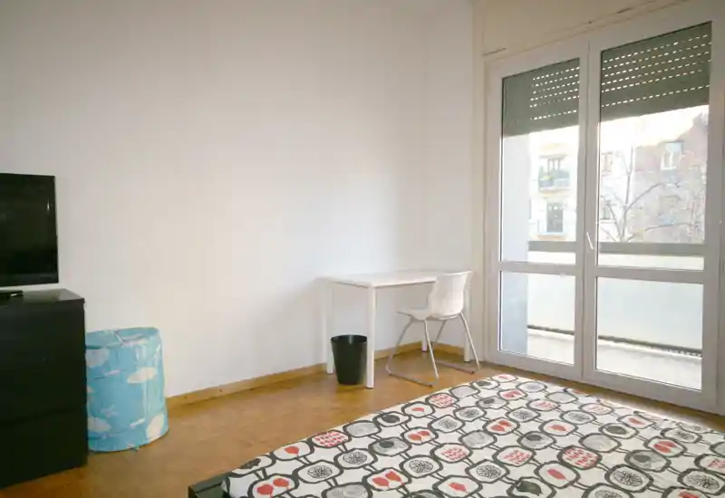 Dormitorio luminoso y de mobiliario sencillo con edredón estampado, un pequeño escritorio junto a una gran ventana y suelo de parquet — ideal para una persona que busca luz natural y un espacio minimalista.