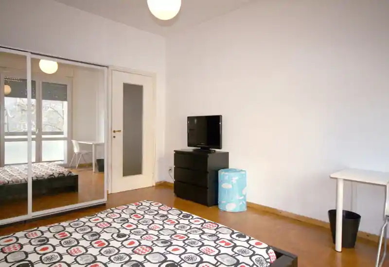 Dormitorio luminoso y minimalista con cama doble estampada, armario con espejo y pequeño escritorio junto a la ventana: líneas limpias y luz natural que aportan calidez.