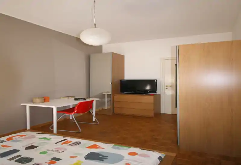 Dormitorio luminoso y amueblado de forma sencilla con cama estampada, escritorio blanco y silla roja, armario y cómoda con TV — líneas claras y acabados en madera.