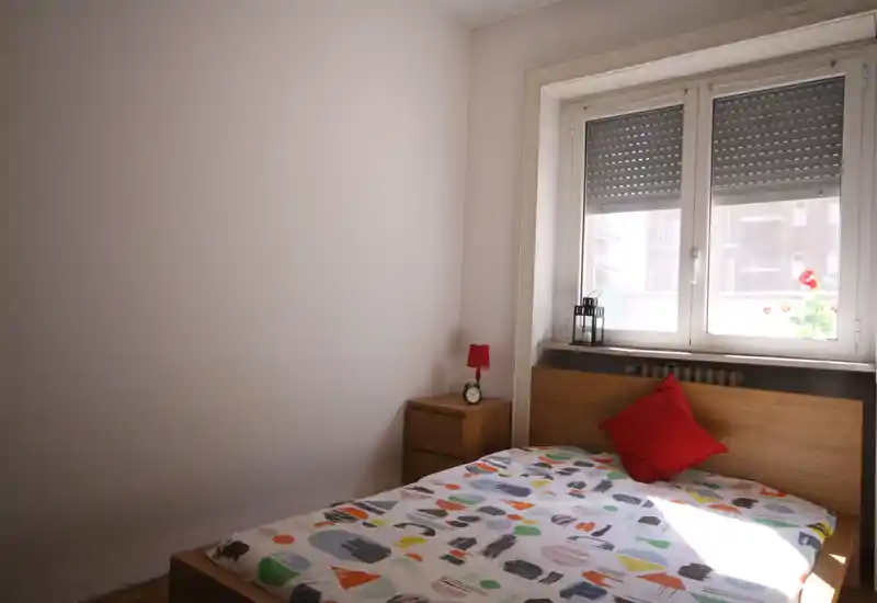 Dormitorio sencillo y luminoso con cama de madera y ropa de cama de estampado colorido. Un cojín rojo y una mesita con lámpara aportan personalidad; luz natural desde la ventana.