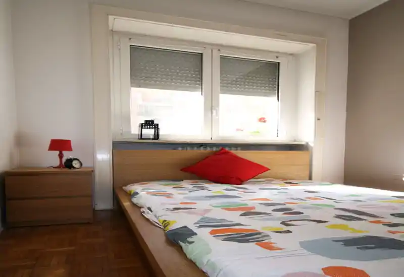 Dormitorio soleado con cama de madera, edredón colorido y un cojín rojo como acento; mesita de noche con lámpara y despertador. La ventana grande aporta luz natural y un aspecto moderno y limpio.