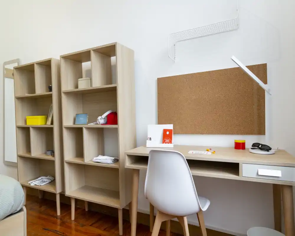 Angolo di camera luminoso e ordinato con scaffalature in legno aperte e una scrivania compatta ideale per studio o lavoro da remoto. Decorazioni sobrie e pavimento in legno creano un ambiente tranquillo e funzionale.