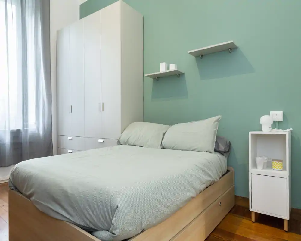 Camera da letto moderna e luminosa con letto matrimoniale, biancheria verde chiaro e parete accentata verde. Armadio bianco, mensole e pavimento in legno naturale creano un ambiente accogliente.