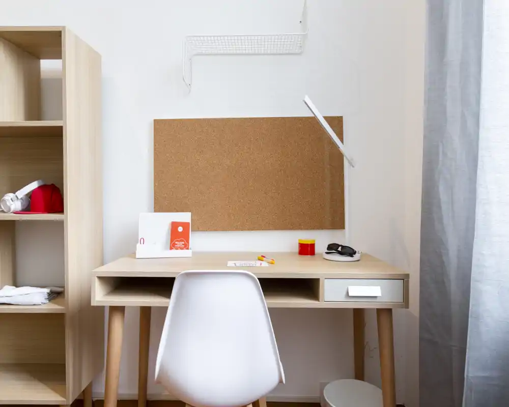 Zona studio luminosa e minimal con bacheca in sughero, lampada e scaffalatura semplice — un angolo lavoro ordinato integrato nell'ambiente abitativo.