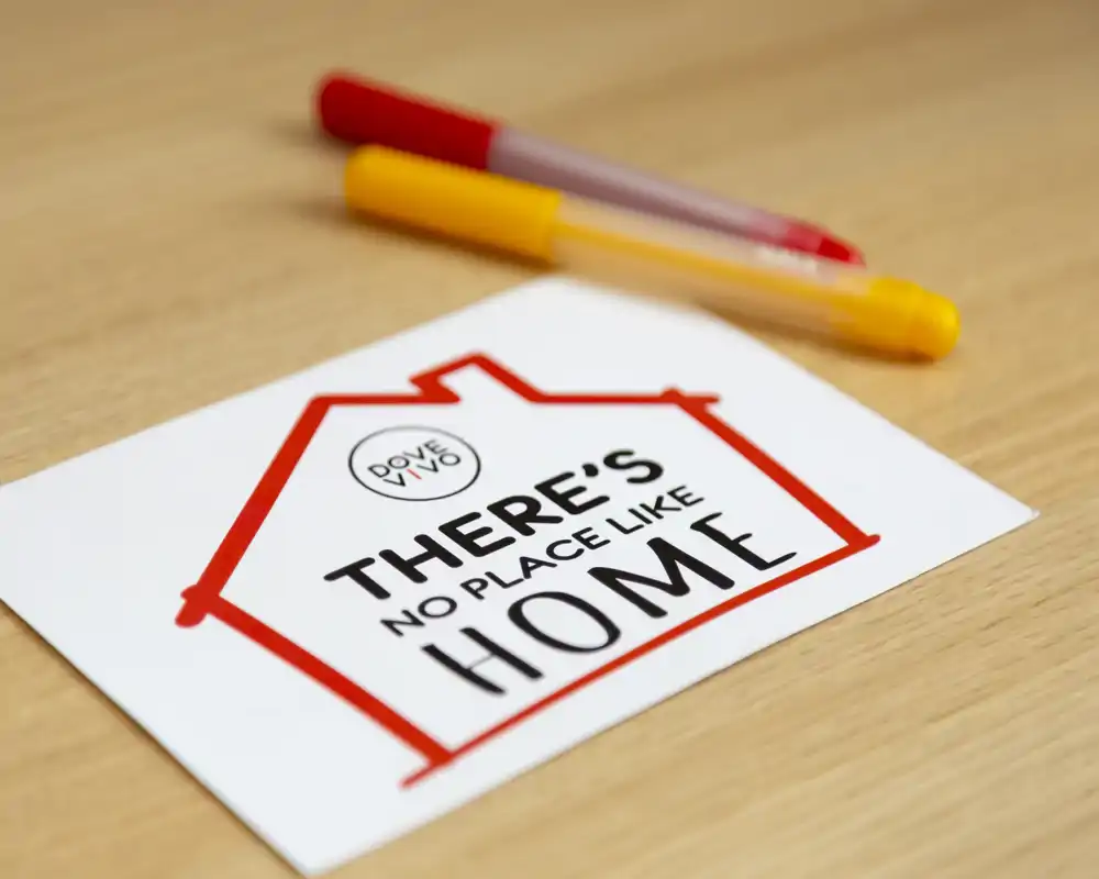 Primo piano di una card su un tavolo in legno con la scritta "There's no place like home" e due penne. Scatto di dettaglio/oggetto, non una foto che mostri una stanza interna.