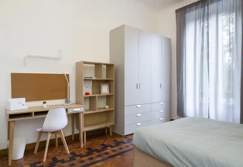 Camera da letto luminosa e ordinata con letto a destra, scrivania e scaffalatura a sinistra e un armadio alto vicino a una finestra che lascia entrare luce naturale.