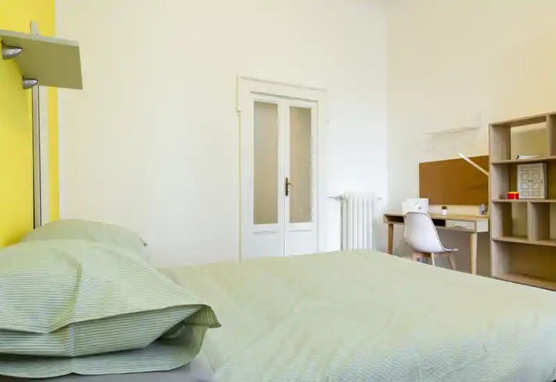 Camera da letto luminosa e ordinata con un ampio letto con biancheria verde tenue, una scrivania compatta e scaffalatura — ideale per un soggiorno confortevole o per studenti.