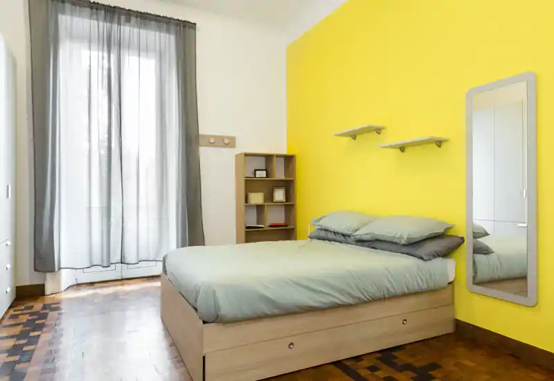 Camera da letto luminosa e moderna con letto matrimoniale, biancheria morbida e una parete gialla vivace. La luce naturale entra dalle tende alte; specchio a figura intera e scaffalatura offrono praticità.