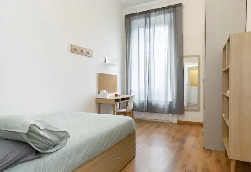 Camera da letto luminosa e minimale con letto singolo, pavimento in legno e una piccola scrivania davanti a una finestra alta con tenda semitrasparente. L'ambiente è pulito, moderno e accogliente, ideale per uno studente o una persona sola.
