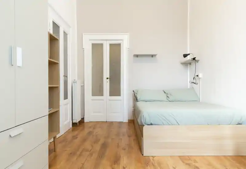 Camera da letto luminosa e minimal con letto matrimoniale, pavimento in legno chiaro e biancheria pastello; linee pulite e mensole semplici creano un'atmosfera tranquilla e ariosa, perfetta per annunci.