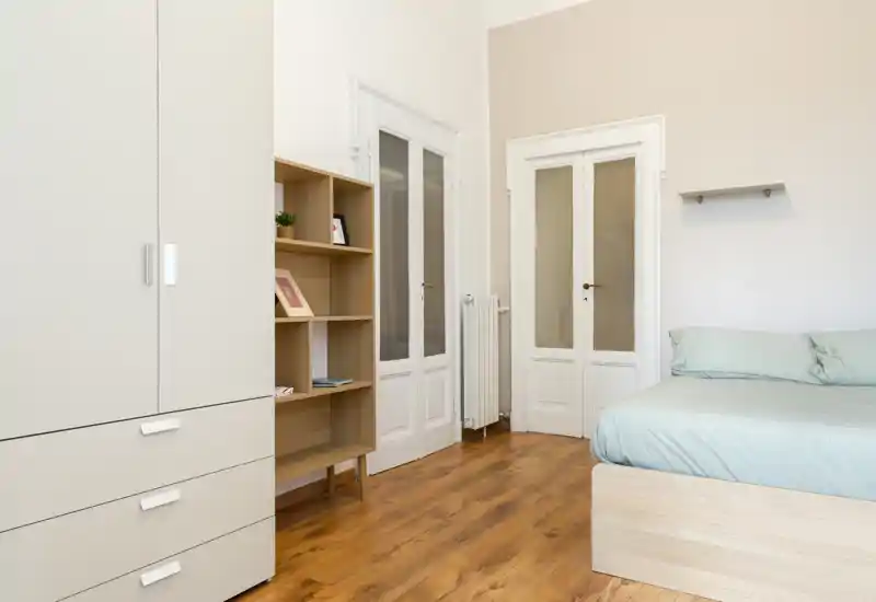 Camera da letto luminosa e minimal con letto in legno chiaro e biancheria pastello, armadio e scaffalatura aperta; pavimento in legno caldo e pareti neutre creano un'atmosfera calma e accogliente.