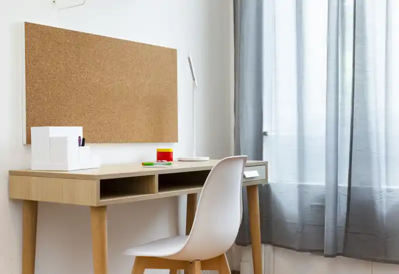Angolo studio luminoso e minimal con scrivania in legno, sedia bianca e bacheca in sughero accanto alla finestra con tenda leggera.