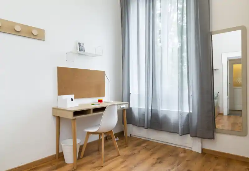 Angolo di camera da letto luminoso e minimal con scrivania in legno, sedia e specchio a figura intera accanto a una grande finestra con tenda.