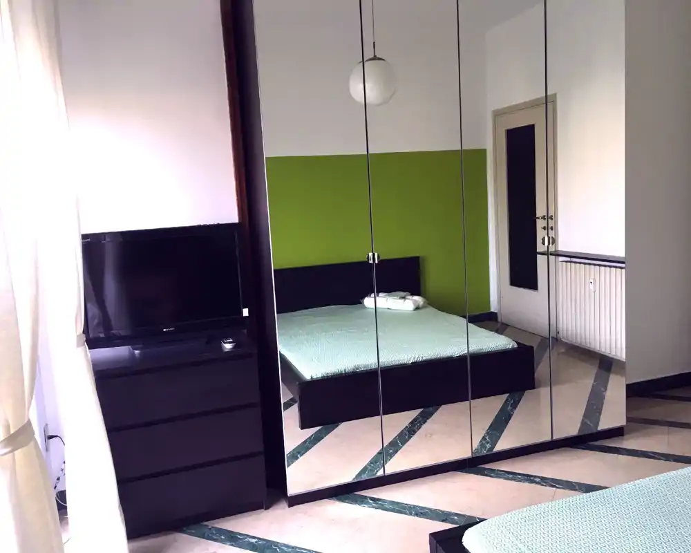 Bright, modern bedroom with a double bed facing mirrored wardrobes; natural light from a window and a simple TV cabinet complete the layout.