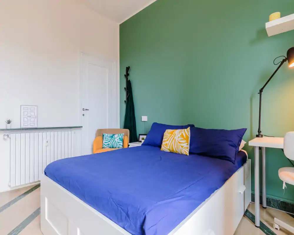 Bright, modern bedroom with a full-size bed dressed in blue linens against a green accent wall; includes a bedside desk, armchair and a floor lamp — tidy and well staged for rental photos.