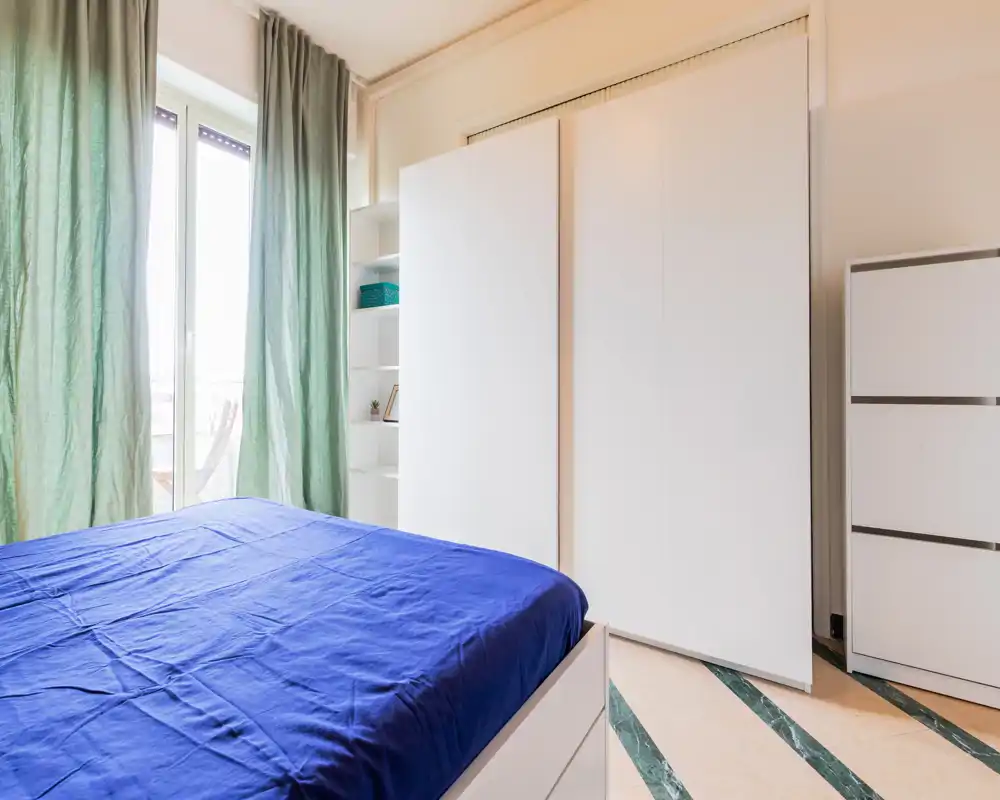 Bright, modern bedroom with a blue-dressed bed, large wardrobe and floor-to-ceiling window letting in ample natural light.