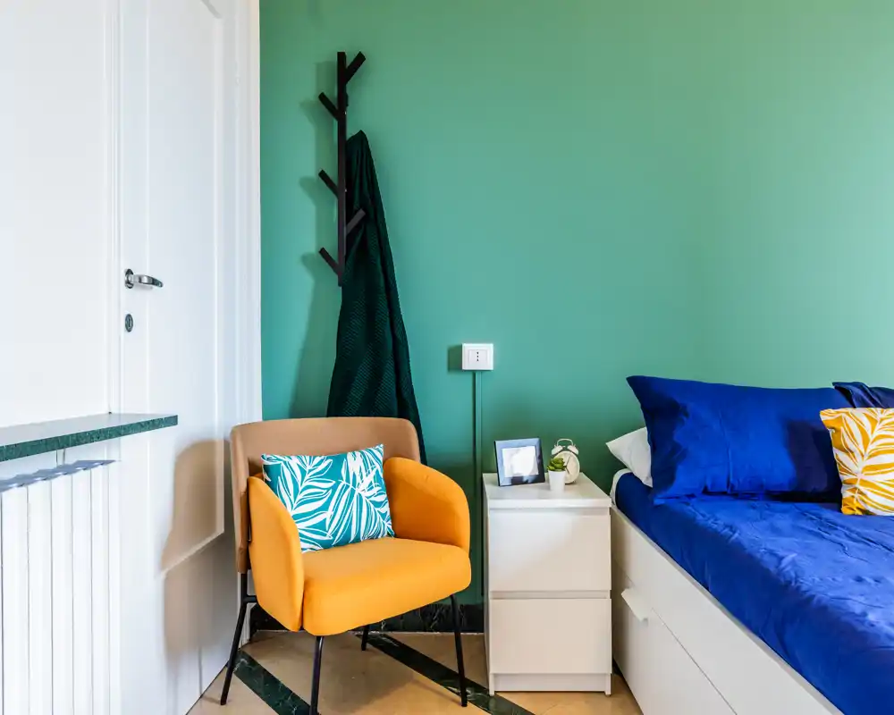 Bright, stylish bedroom corner with a blue-dressed bed, white bedside table and a warm mustard armchair against a green wall — modern, cozy and well-lit.