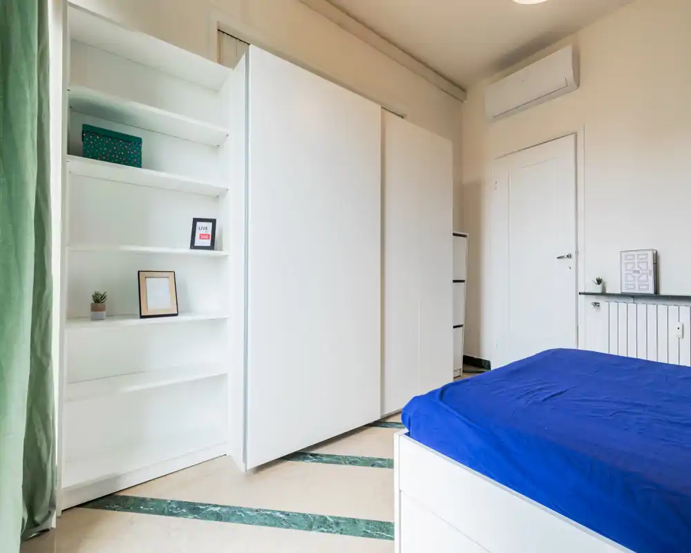Bright, modern bedroom with a sizable white wardrobe, simple shelving and a bed with deep blue bedding. Clean, minimal decor and good natural light make this room inviting for rental photos.