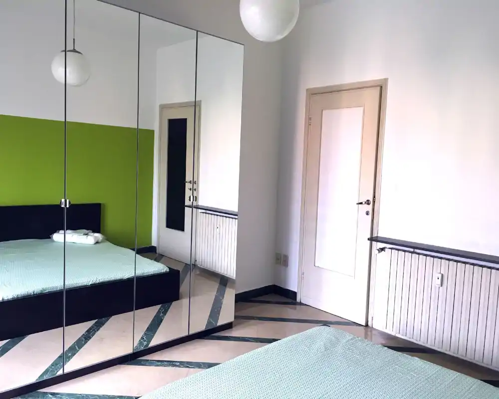 Bright, minimalist bedroom with a large mirrored wardrobe reflecting the bed; clean lines and soft green accent wall create a calm atmosphere.