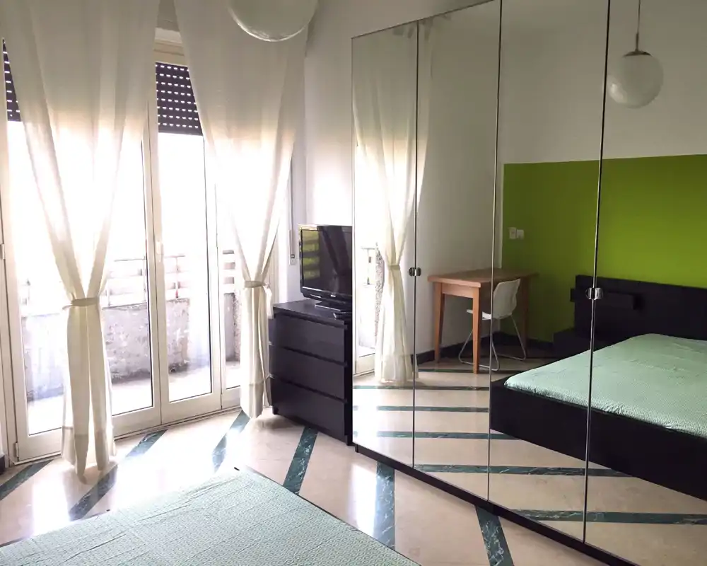 Bright, modern bedroom with a large bed, mirrored wardrobe and balcony doors that flood the room with natural light. Simple desk and dresser provide practical workspace and storage.