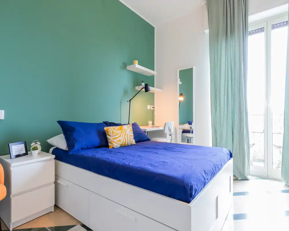 Bright, modern bedroom with a blue bed against a green accent wall, bedside table, desk with lamp and large window providing abundant natural light.