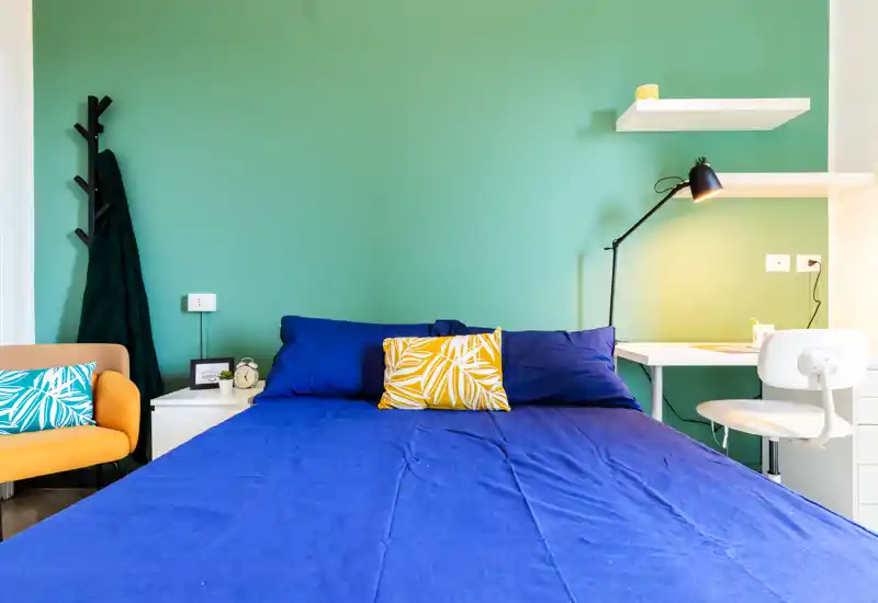 Bright, modern bedroom with a large blue bed, decorative cushions and a small workspace at the side—fresh green wall and tidy styling ideal for rental photos.