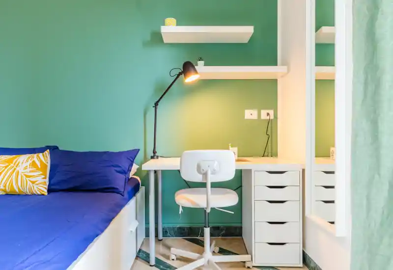Bright, modern bedroom corner with a neatly made single bed, blue linens and a yellow decorative cushion; a white desk and chair with a reading lamp make a functional workspace against a green wall.
