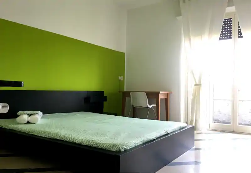 Bright, modern bedroom with a low platform bed against a bold green accent wall and a small desk by the balcony door letting in abundant natural light.