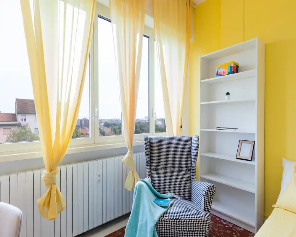 Rincón de dormitorio luminoso y alegre con grandes ventanas, cortinas amarillas, un sillón estampado y una estantería blanca — perfecto para un anuncio de alquiler soleado.