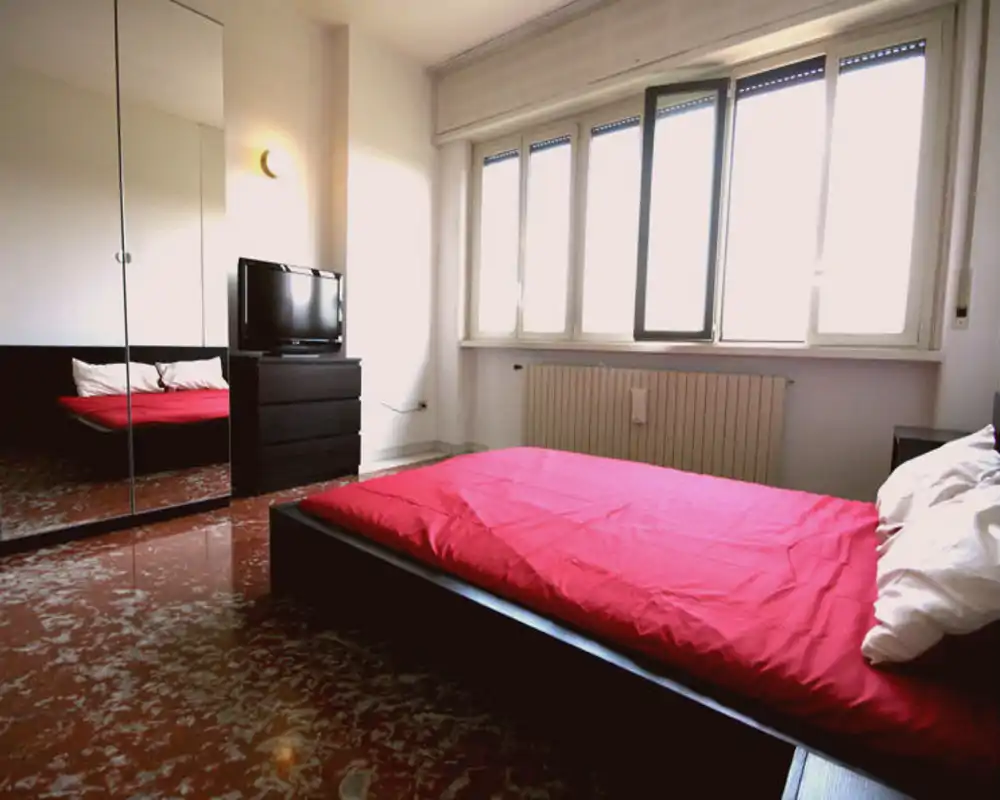 Dormitorio moderno y luminoso con cama baja y edredón rojo, gran ventana que aporta luz natural, armario con espejos y cómoda con televisor.