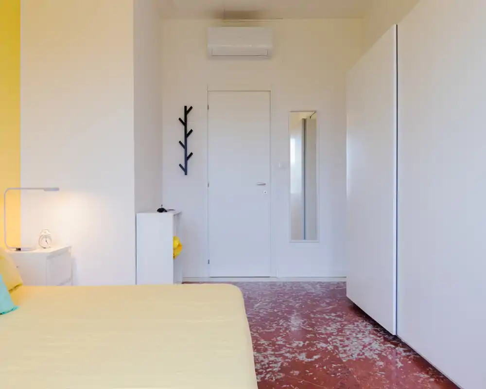 Dormitorio minimalista y limpio con colcha amarillo claro, lámpara de mesilla y armario; espacio luminoso y bien presentado, ideal para el anuncio.