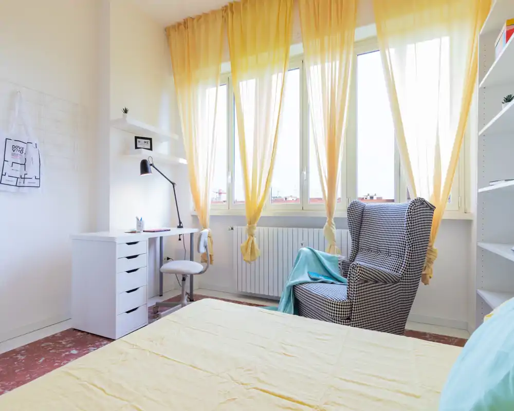 Dormitorio luminoso y aireado con grandes ventanas, cama en primer plano, un sillón estampado y un pequeño escritorio blanco; cortinas amarillas que filtran la luz natural para una atmósfera acogedora.