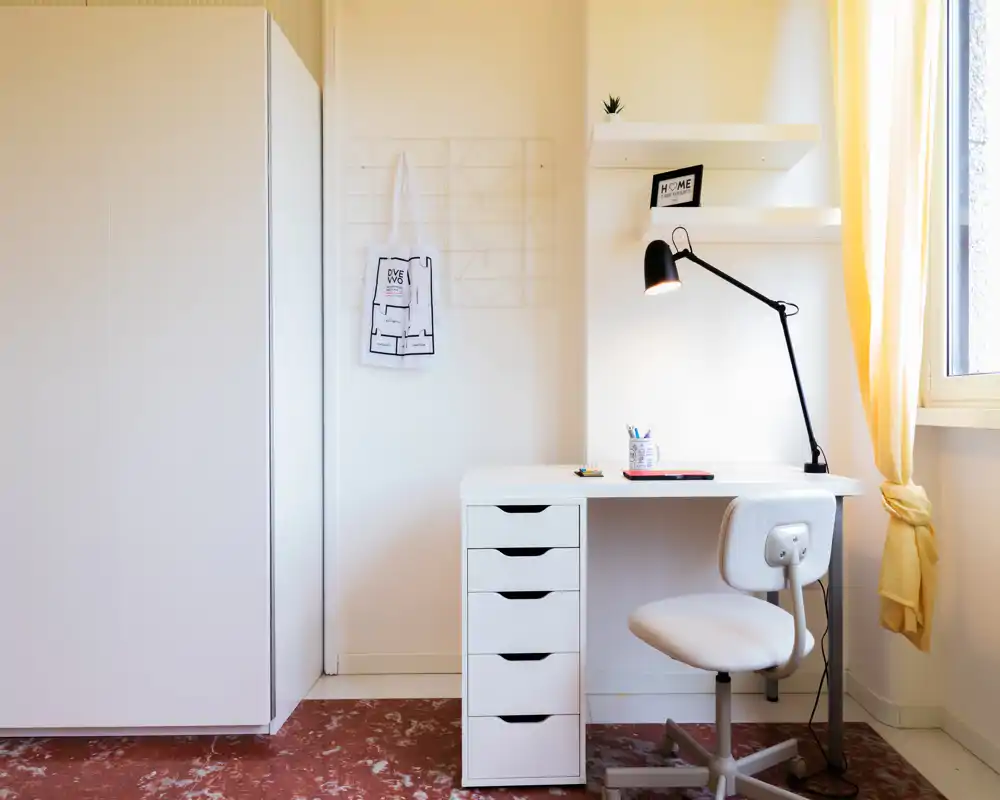 Rincón de dormitorio luminoso y minimalista con escritorio blanco, estantes y una silla giratoria junto a una ventana con cortinas amarillas. Líneas limpias, luz natural cálida y decoración sencilla crean un espacio de trabajo tranquilo.