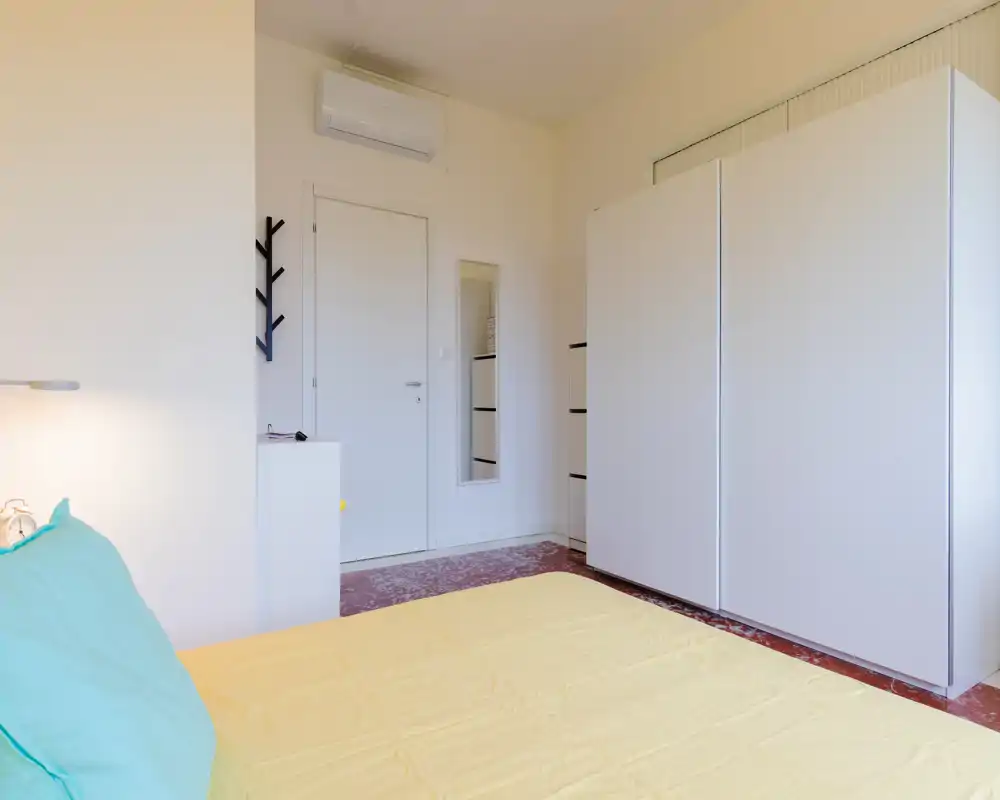 Dormitorio luminoso y ordenado con la cama en primer plano, un gran armario blanco y un espejo de cuerpo entero junto a la puerta.