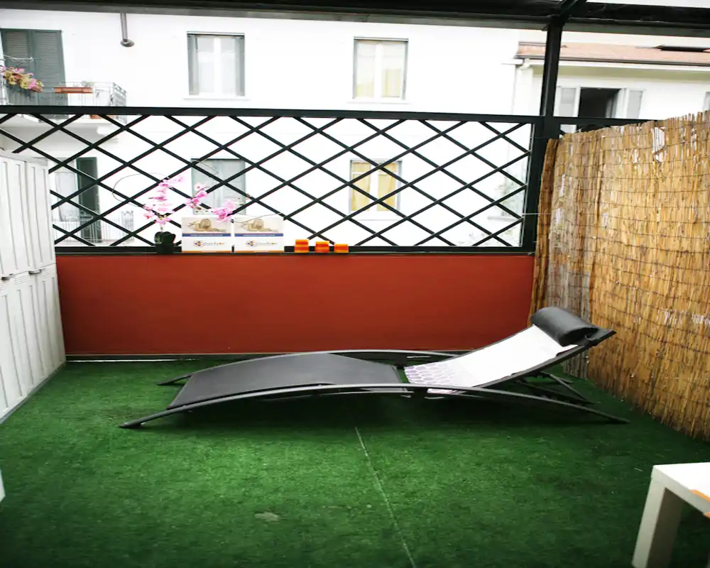 Balcón cubierto y soleado con tumbona reclinable, suelo de césped artificial y pantalla de cañas para privacidad — un rincón exterior acogedor para relajarse.