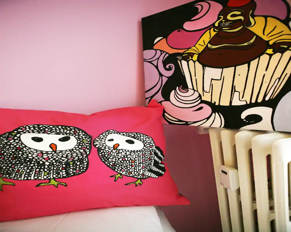 Primer plano de un rincón del dormitorio con una almohada decorativa de búhos en la cama, un cuadro colorido y un radiador: una toma acogedora y divertida del área de descanso.