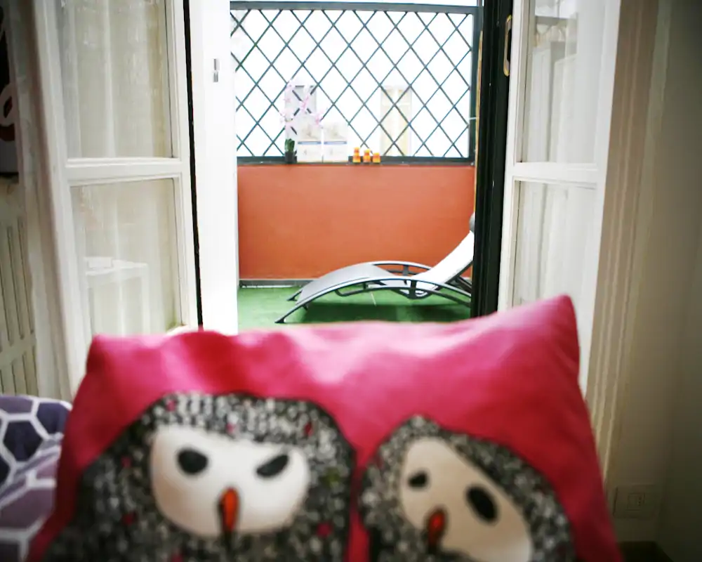 Pequeña terraza soleada con una tumbona visible a través de puertas balconeras; un cojín decorativo en primer plano añade calidez y profundidad.