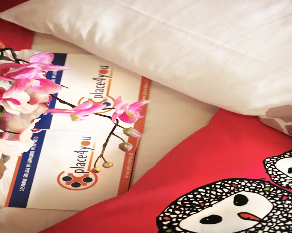 Primer plano de la esquina de una cama con almohada blanca, cojín decorativo y una pequeña orquídea sobre tarjetas promocionales; imagen luminosa y acogedora, ideal para destacar ropa de cama y decoración.