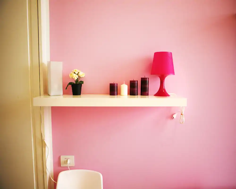 Un rincón de sala minimalista y con estilo: una repisa flotante con velas, una pequeña planta y una lámpara rosa frente a una pared rosa pastel.