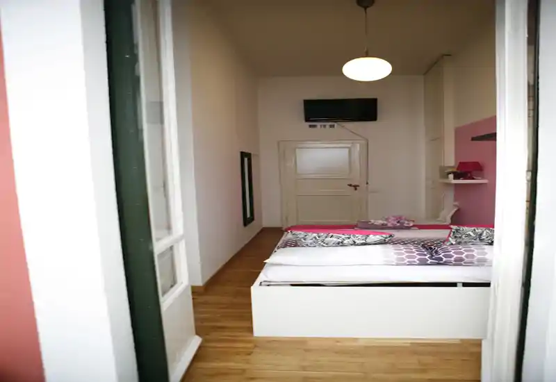 Bright, well-styled bedroom with a double bed, patterned bedding and a pink accent wall; wooden floor and mounted TV complete the simple, welcoming layout.
