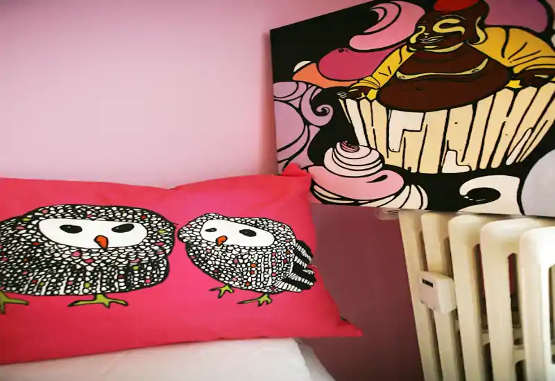 Close-up of a bedroom corner showing a decorative owl pillow on the bed, colorful wall art and a radiator — a cozy, playful detail shot of the sleeping area.