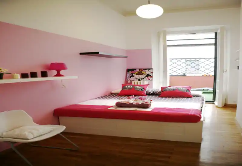 Bright, cozy bedroom with a neatly made double bed, decorative cushions and soft pink accent wall opening onto a small balcony.