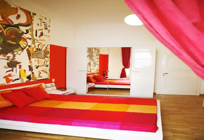 Bright, colorful bedroom with a large low platform bed, vibrant red and orange linens, mirrored wardrobe and decorative wall art — a warm, stylish space ideal for rental listings.